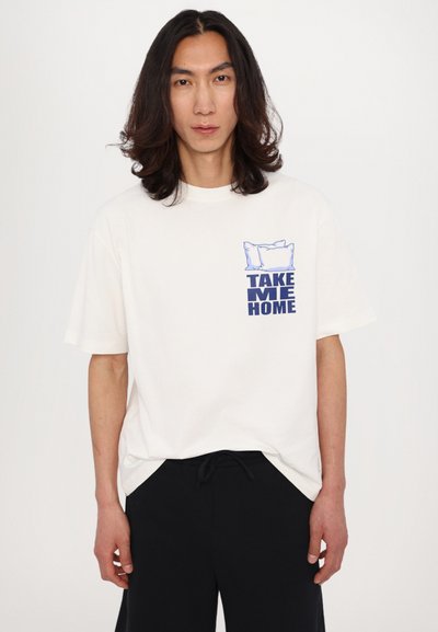 Witte oversized T-shirt met korte mouwen, voorzien van een blauwe grafische afbeelding van kussens en de tekst "NEEM ME MEE NAAR HUIS" op de linkerkant van de borst.
