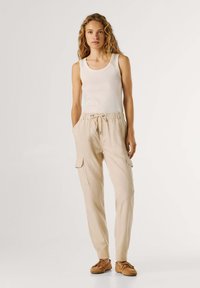 Młoda kobieta o kręconych włosach, ubrana w biały bezrękawnik, beżowe spodnie typu cargo z troczkiem w talii oraz brązowe casualowe buty, stojąca na tle białego tła.