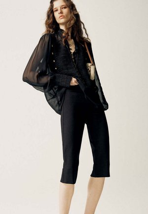 Femme portant un chemisier noir transparent à manches bouffantes, pantalon noir court, tenant un petit sac à épaule sur un fond neutre.