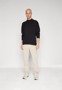 Suéter de cuello alto negro, pantalones de pana beige y zapatillas grises. Textura suave y corte relajado, con mangas remangadas y un estilo casual.