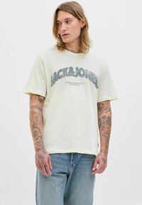 Šviesiai geltona medvilninė trumpomis rankovėmis marškinėliai, su tamsiai mėlynu grafiku "JACK&JONES". Dėvima su mėlynomis džinsinėmis kelnėmis.