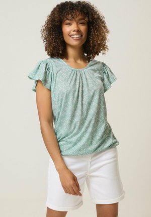 Lachende vrouw met krullend haar, gekleed in een mintgroene blouse met korte mouwen en bloemenprint, en witte opgerolde korte broek, staand voor een effen achtergrond.