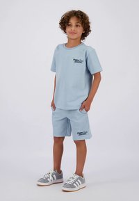 Camiseta de punto de manga corta en azul claro con pantalones cortos a juego. Presenta una tela texturizada y un pequeño logo impreso en el pecho. Zapatillas deportivas grises.