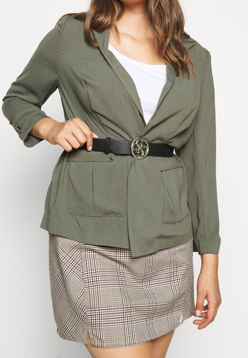 Femme portant un blazer vert olive avec une ceinture noire et une jupe à carreaux, ajustant la ceinture d'une main.