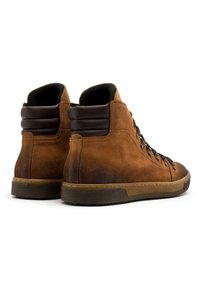 Brązowe zamszowe buty wysokie z ciemnymi skórzanymi akcentami, zaokrąglony nosek, sznurowany design oraz teksturowana gumowa podeszwa. Widok z tyłu pokazuje wyściełany kołnierz.