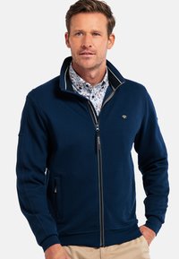 Navy zip-up jacket in textured fabric with a high collar, featuring a logo on the chest and side pockets. Paired with a floral-patterned shirt.