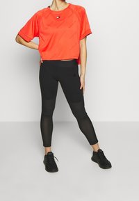 Czerwony sportowy krótki top z krótkimi rękawami i wzorem logo, w zestawie z czarnymi legginsami z siateczkowymi panelami i logo. Czarne buty sportowe.