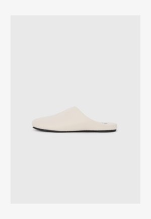 Beige slip-on slipper with a soft, textured upper and black rubber sole. Features a minimalist design with a low profile and no visible hardware.