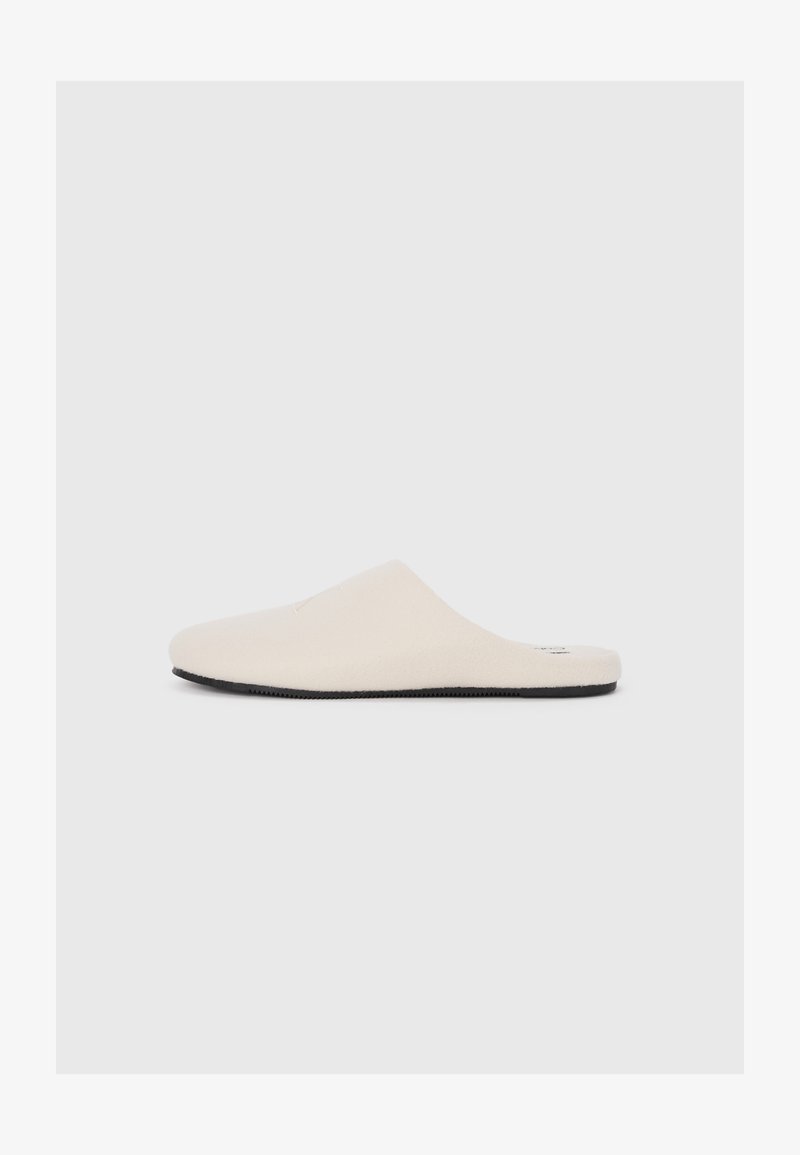 Beige tøfler med mykt, teksturert overmateriale og svart gummisåle. Har et minimalistisk design med lav profil og ingen synlige feste.