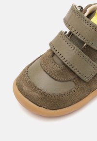 Olive zaļas krāsas mazuļa kurpes, izgatavotas no zamša un ādas materiāliem, ar divām Velcro siksnām un gumijas zoli. Noapaļots purngals un šūšanas akcenti.