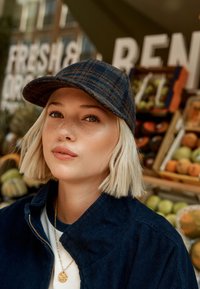 Die Mütze verfügt über ein dunkelblaues und braunes Karomuster und besteht aus einer Wollmischung. Das Modell trägt eine marineblaue Cordjacke mit einem weißen Hemd darunter.