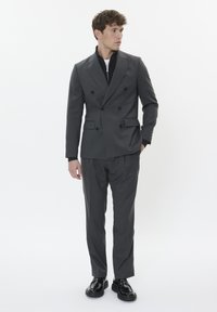 Hall hallitusega hall topeltpükste ülikond, millel on kergelt kohandatud fits, varustatud nööbiga kraega ja küljetaskutega, kombineeritud läikivate mustade kingadega.