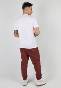 T-shirt blanc à manche courte associé à un pantalon de jogging bordeaux avec des accents brodés, des chevilles retroussées et des baskets blanches sur un fond gris clair.