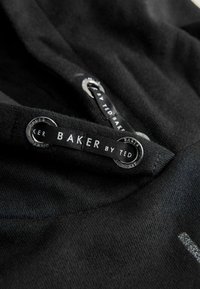 Černá tkanina s hladkým povrchem má výrazný kruhový stříbrný kovový detail a černou tkaninovou visačku s nápisem "BAKER by TED."