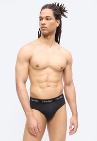 Izmos férfi dreadlock hajjal, fekete Calvin Klein alsónadrágban, balra nézve, egyszerű világos háttér előtt.