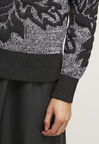 Sweter dzianinowy w czarno-białe wzory, z prążkowanymi mankietami i dolnym wykończeniem, charakteryzujący się wyraźną teksturą i abstrakcyjnym wzorem.