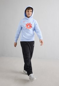 Chlapec chodí uvnitř a má na sobě světle modrou mikinu s kapucí a oranžovým logem, černé sportovní kalhoty Nike a bílé tenisky, v pozadí je jednoduchá šedá stěna.