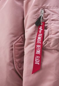 Porte-clés en tissu rouge avec texte blanc "Remove Before Flight" attaché à la poche zippée d'une veste rose.