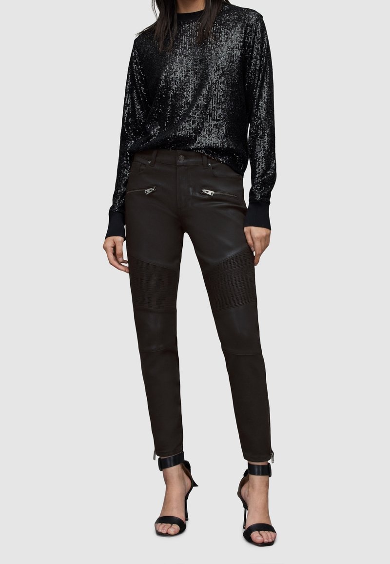 Haut noir à manches longues orné de sequins, associé à un pantalon en cuir noir ajusté avec des zips et des détails côtelés, complété par des sandales noires à talons hauts.