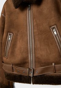 Veste en peau de mouton brune avec une fermeture éclair, des accents en cuir, un col en polaire et une ceinture ajustable à la taille. Elle dispose de deux poches zippées.