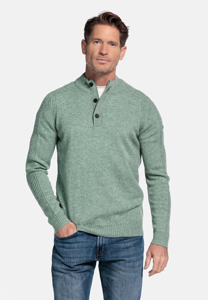 Lichtgroene gebreide trui met een hoge kraag en drie donkere knopen. Geribbelde manchetten en zoom, gedragen over een witte t-shirt, gecombineerd met blauwe jeans.