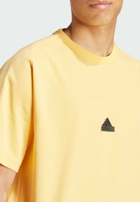 Żółta koszulka krótki rękaw z bawełny, z okrągłym dekoltem i czarnym logo Adidasa na piersi. Miękka tekstura, luźny krój.