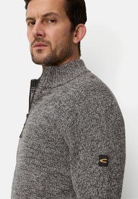 Mand med stubbe, der bærer en grå strikket lynlåstrøje med en sort og orange logo på øverste ærme, kigger til siden.