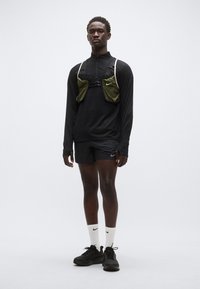 Top preto de mangas compridas, mochila de hidratação verde com dois bolsos e alças ajustáveis, calções pretos, meias brancas e ténis desportivos pretos.