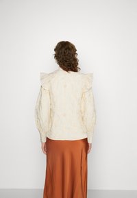 Blusa bianca ricamata con fiori, con spalle arricciate, maniche lunghe, tessuto testurizzato, abbinata a una gonna in satin marrone.