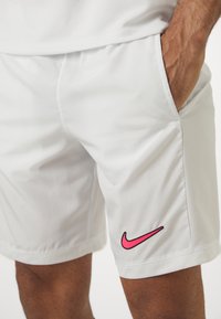 Fehér sportnadrág sima textúrával és légáteresztő anyaggal. A bal combon található egy kis fekete és rózsaszín Nike logó.