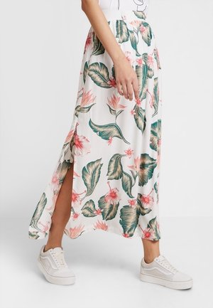 Jupe longue fleurie en tissu léger, avec de grandes feuilles vertes et des fleurs roses. Conception avec fente latérale. Associée à des baskets blanches.