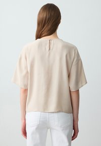 Beige korte mouwen top gemaakt van lichte stof, met een ronde halslijn en een sleutelgaatje op de rug. Draagt met witte broek.