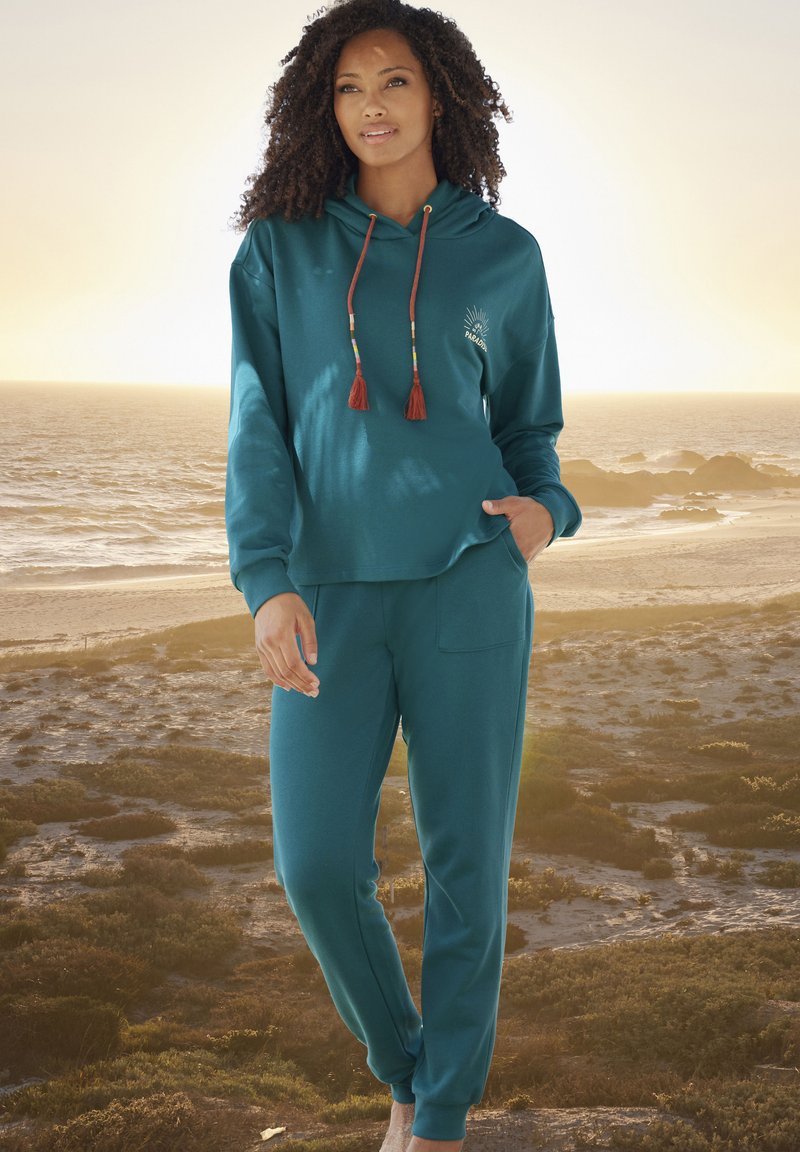 Felpa teal con frange decorative e logo, abbinata a pantaloni coordinati. Tessuto morbido, vestibilità comoda e tasche frontali per funzionalità.