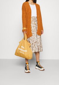 Pomarańczowy, puszysty kardigan, biały top, spódnica midi w cętki, żółta torba na ramię z napisem oraz czarno-białe trampki z pomarańczowymi akcentami.