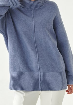 Personne portant un pull en tricot bleu ample à col roulé avec des poignets et un ourlet côtelés, associé à un pantalon blanc, sur un fond uni.