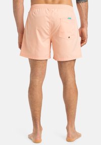 Shorts de bain pêche avec une taille élastique, une poche arrière et une petite étiquette de marque. Tissu lisse avec un design arrivant au milieu de la cuisse.
