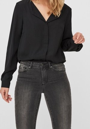 Woman wearing a black long-sleeve blouse tucked into high-waisted faded black skinny jeans.