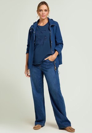 Donna con giacca blu, camicia blu navy ricamata, jeans a gamba larga e mocassini color cuoio, in piedi con una mano in tasca.