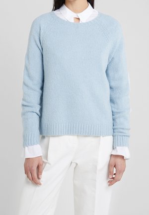 Osoba ubrana w jasnoniebieski sweter z dzianiny na białą koszulę z kołnierzykiem, w białych spodniach, na jednolitym szarym tle.