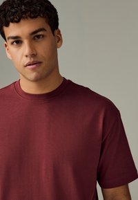 Bordové tričko s okrúhlym výstrihom a krátkymi rukávmi, vyrobené z mäkkej látky. Dizajn zdôrazňuje jednoduchý, čistý strih a detailné textúrované švy.