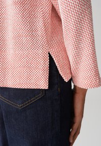 Texturiertes Oberteil aus Korallen- und weißem Stoff mit seitlichem Schlitz und Dreiviertelärmeln, kombiniert mit dunkelblauen Jeans.
