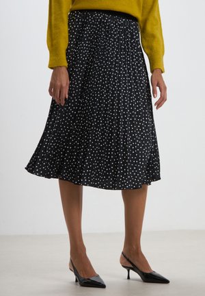Gonna nera plissettata con pois bianchi, design a lunghezza media, tessuto leggero, abbinata a decolté nere a punta, indossata con un maglione giallo.