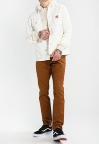 Witte katoenen jas met een zachte textuur en een patchzak, over een beige fleece hoodie, gecombineerd met bruine broek en zwarte sneakers.