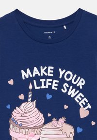 Tume sinine T-särk, millel on cupcake'i ja koogi illustratsioonid ning tekst "TEE OMA ELUST MAGUS" roosade ja siniste südamekestega ümbritsetuna.