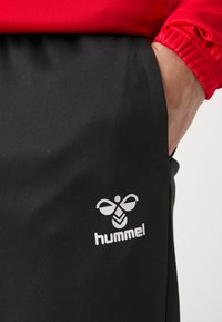 Melni sporta bikses, kas izgatavotas no gludas auduma, ar baltu Hummel logo uz kreisās kājas un sarkanu elastīgu aproci augšā.