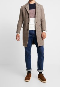 Hombre con abrigo beige a cuadros sobre suéter de colores, jeans azules arremangados, calcetines negros y zapatos de ante marrones sobre fondo blanco.