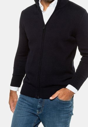 Mand iført sort cardigan med lynlås over hvid skjorte med opsmøgede ærmer, blå jeans, med den ene hånd i lommen, mod hvid baggrund.