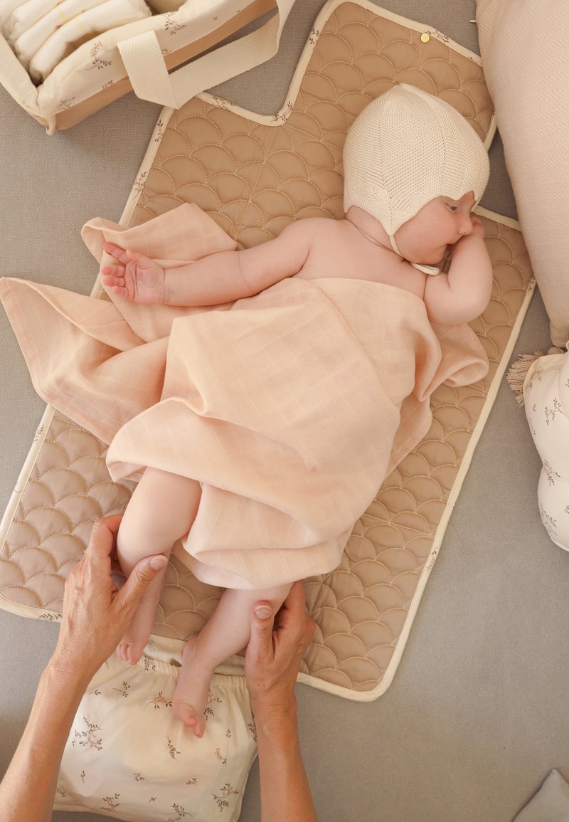 Bambino avvolto in una coperta pesca che indossa un cappello di lana, disteso su un tappetino trapuntato, con mani adulte che tengono delicatamente le gambe del bambino.