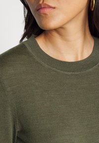 Olivengroene, gestructureerde trui met een ronde halslijn en subtiele geribbelde details. Close-up weergave gericht op de stof en halslijn.