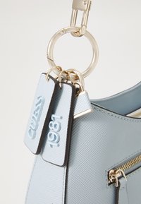 Sac à main synthétique bleu clair avec une finition texturée, quincaillerie dorée, et deux étiquettes estampillées "GUESS" et "1981" attachées à un porte-clés.